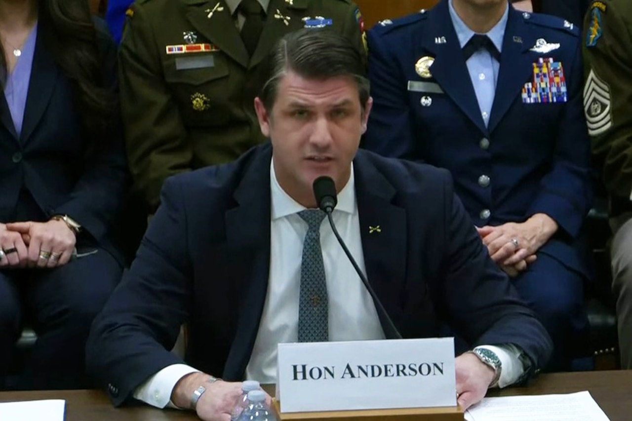A man in a business suit seated at a table with the sign Hon Anderson" and talks into a mic while three people in military uniform and one in civilian attire sit behind him.