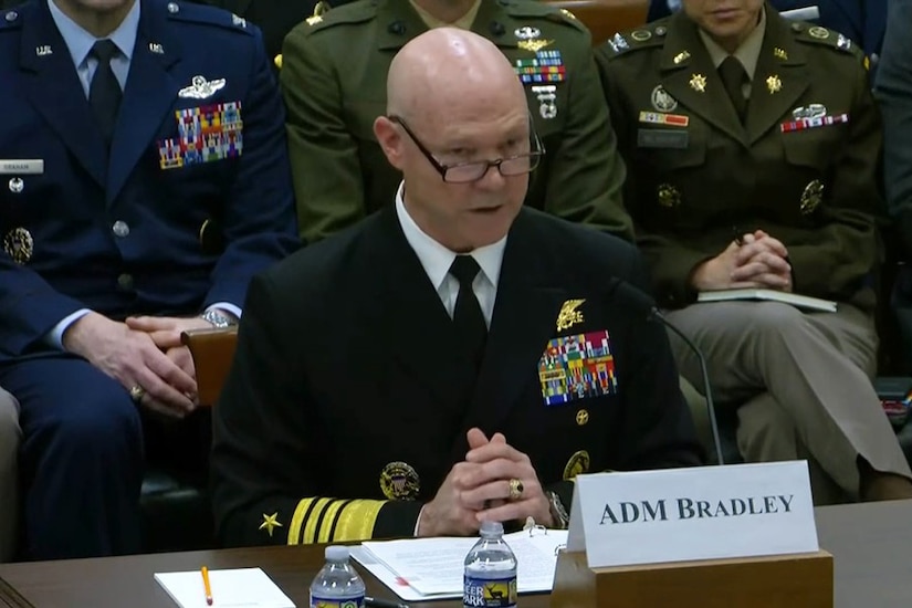 A man in military dress uniform sits at a table with a small sign “ADM Bradley" as he talks into a mic. Three people also in dress uniforms sit behind him.