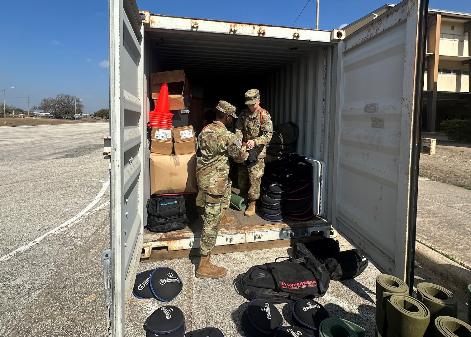 Two people in military uniforms stand in the open door of a shipping container. Property sits on the ground outside of the container.