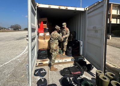 Two people in military uniforms stand in the open door of a shipping container. Property sits on the ground outside of the container.