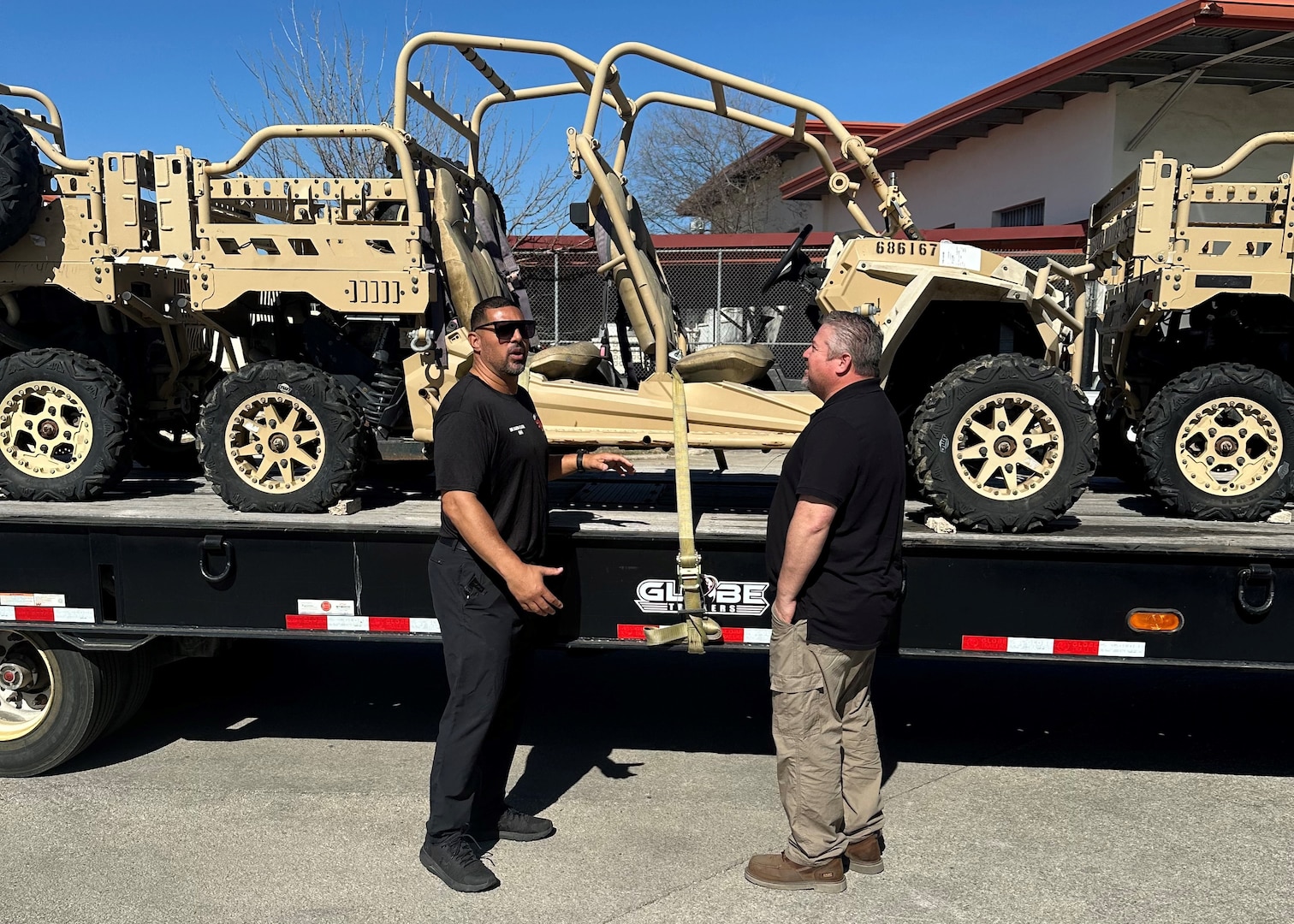two men stand next to a semi truck's flatbed trailer that has tan off road vehicles on it.
