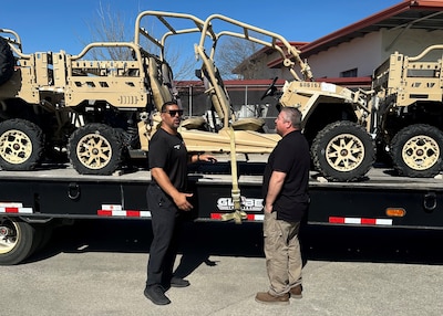 two men stand next to a semi truck's flatbed trailer that has tan off road vehicles on it.