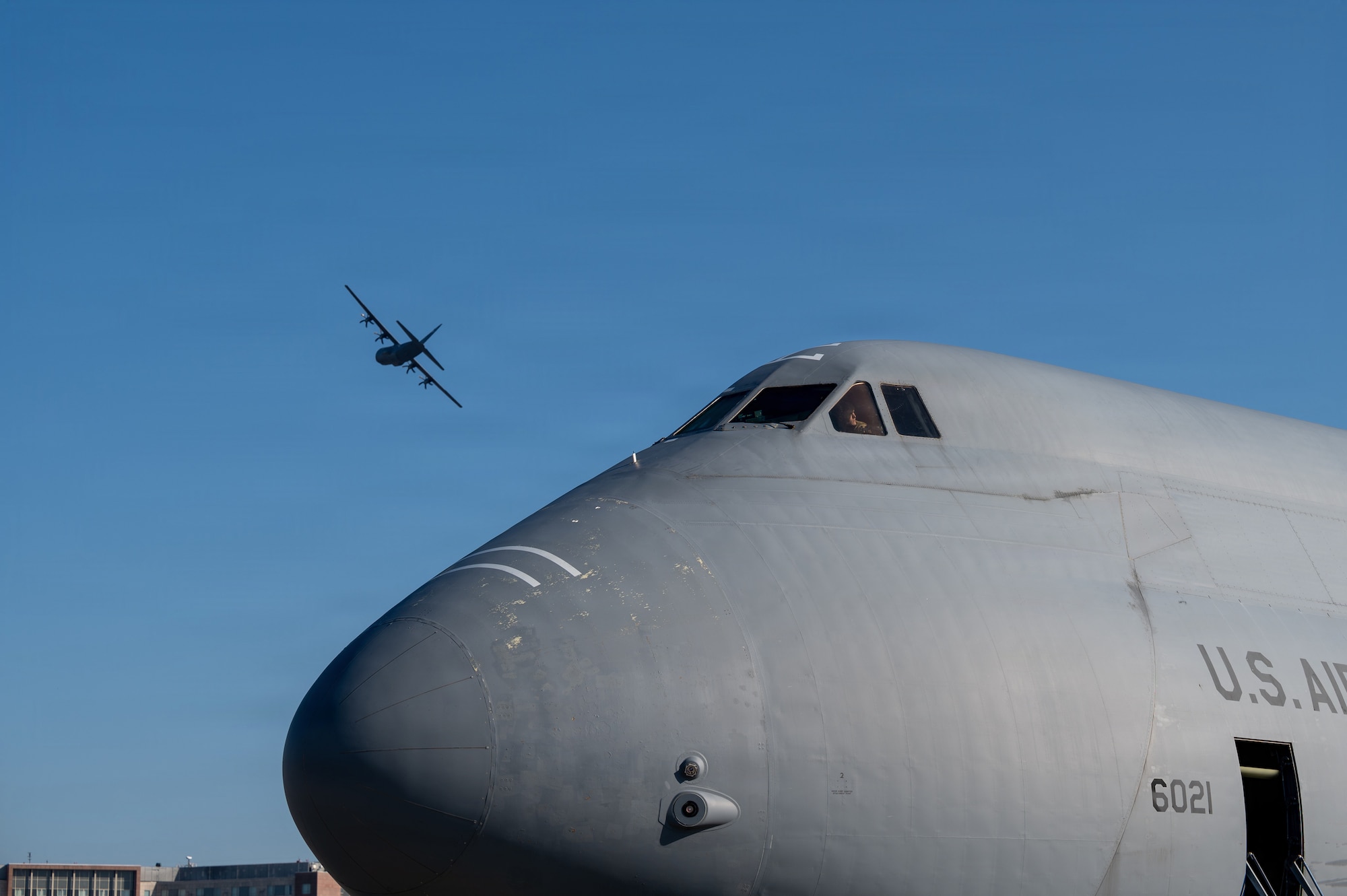 A U.S. Air Force C-5M Super Galaxy sits on the flight line while a C-130 Super Hercules flies overhead at Ramstein Air Base, Germany, March 4, 2026.