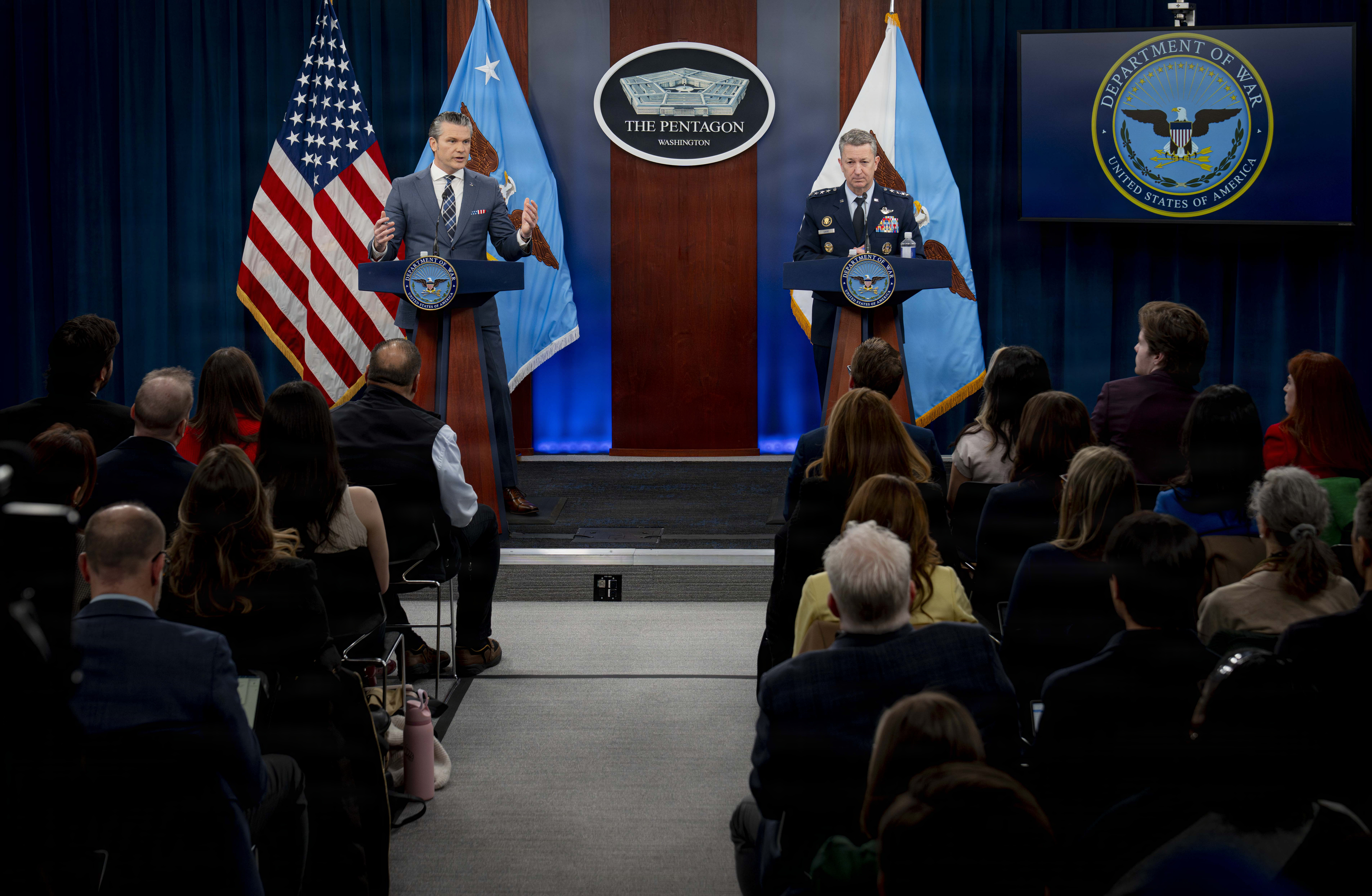 Two men, one in a suit and the other in a military uniform, stand behind lecterns, addressing people. The U.S. flag and two other flags are behind the two men, along with a sign that reads 'The Pentagon.'