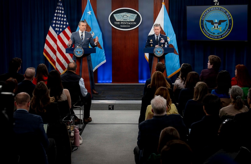 Two men, one in a suit and the other in a dress military uniform, are standing on a stage behind lecterns, addressing a group of people in business attire seated in front of the stage with their backs to the camera. The U.S. flag and the flag of the secretary of war are behind the man in the business suit, and the flag of the chairman of the Joint Chiefs of Staff is behind the man in the military uniform. Between them, mounted on the wall, is a sign that shows a five-sided building that reads "The Pentagon."
