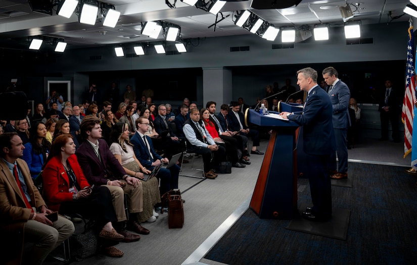Two men, one in a suit and the other in a dress military uniform, are standing on a stage behind lecterns, addressing a group of about 60 people in business attire who are seated and facing the stage. Studio lights are mounted on the ceiling.