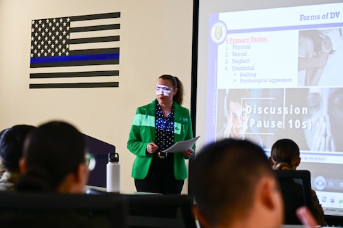 A lady, dressed in a green jacket and dark blouse, is standing in front of a classroom full of Airmen. She is speaking to them.