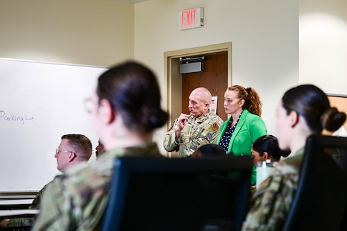 Within a classroom full of airmen, there is an officer and lady standing next to each other. They are both looking ahead, looking closely at what is happening.