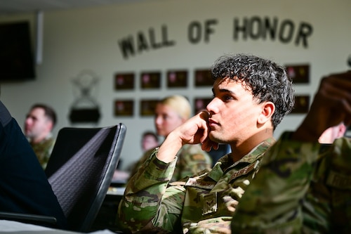 An airman is sitting with his hand propping up his head. He looks to be paying attention very closely to what is happening in front of him. It is a dark lit room with sun hitting his face.