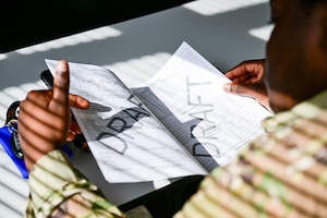 An airman is inspecting papers that looks to be a  checklist and diagram. The word "draft" is written across both pages. There is sunlight peeking through the blinds of a dark room.