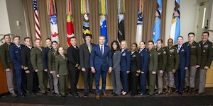 A man wearing a business suit smiles and stands in the middle of a group of people, two of whom are wearing civilian attire and the others in various military dress uniforms while posing for a photo. There is a line of flags behind them.