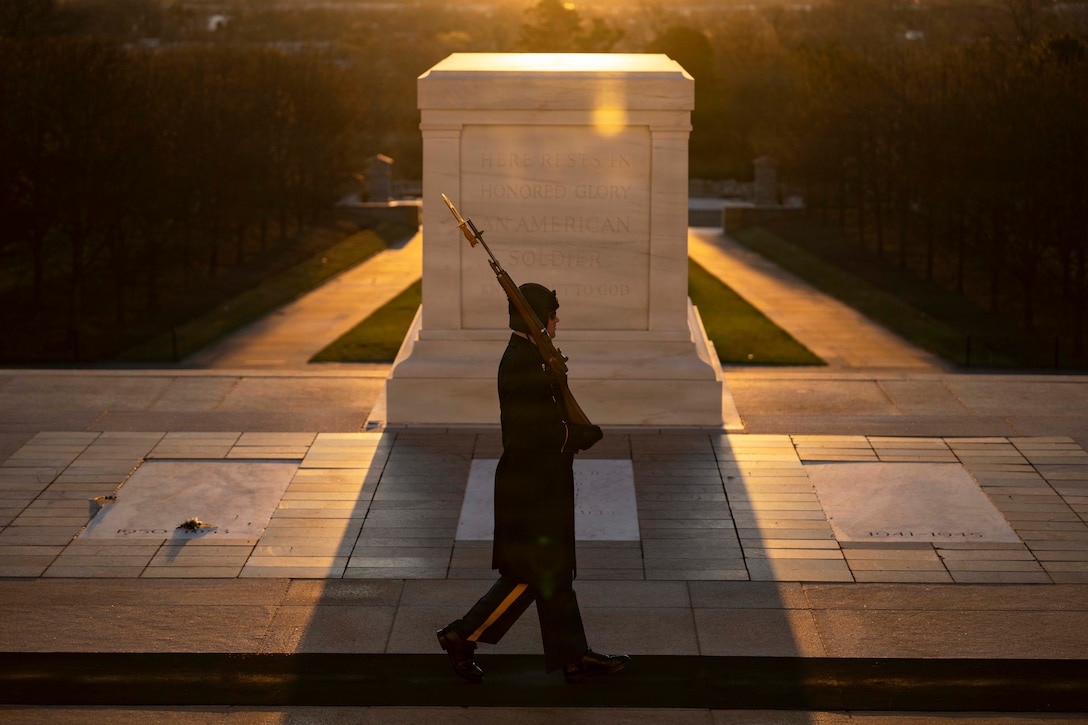 A service member in ceremonial dress carries a rifle while walking in front of a large tomb as the sun rises.