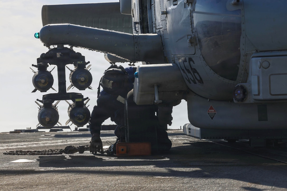 A sailor kneels on the flight deck of a ship to attach chains to a partially visible helicopter during the day.