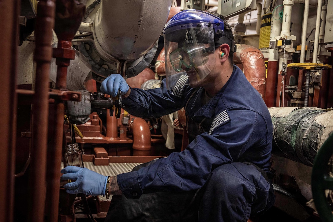 A sailor wearing protective face and hand gear gets a fuel sample from a tank in a maintenance room aboard a ship.