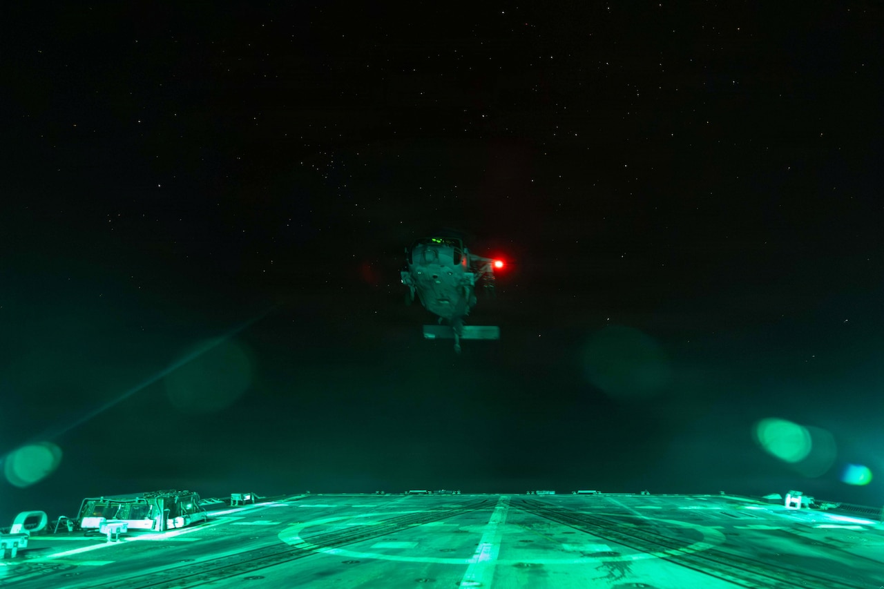 A helicopter illuminated by a red light takes off into a dark sky from the flight deck of a ship.