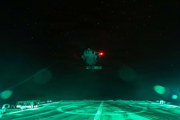 A helicopter illuminated by a red light takes off into a dark sky from the flight deck of a ship.