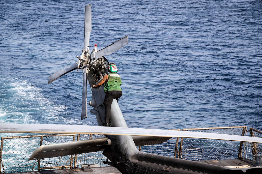 A sailor in a green vest climbs a helicopter to work near the tail rotor aboard a ship at sea.