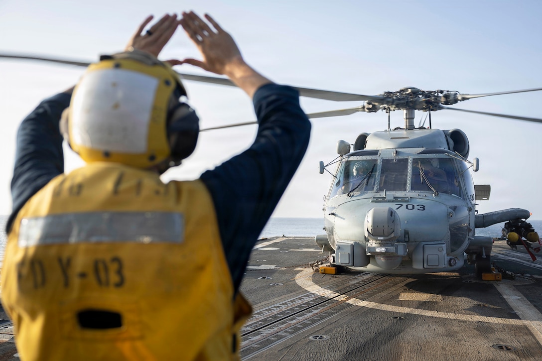 A sailor in a yellow vest forms a triangle with their hands toward a helicopter aboard a ship at sea.