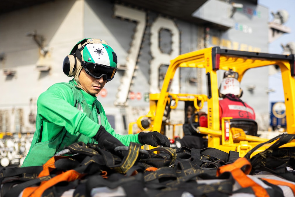 A sailor in a green vest and helmet removes net from a pallet aboard a ship during the day.