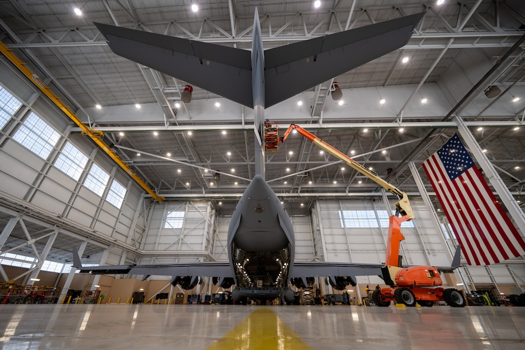 A C-17 is shown in a hangar from behind with an American Flag shown as well