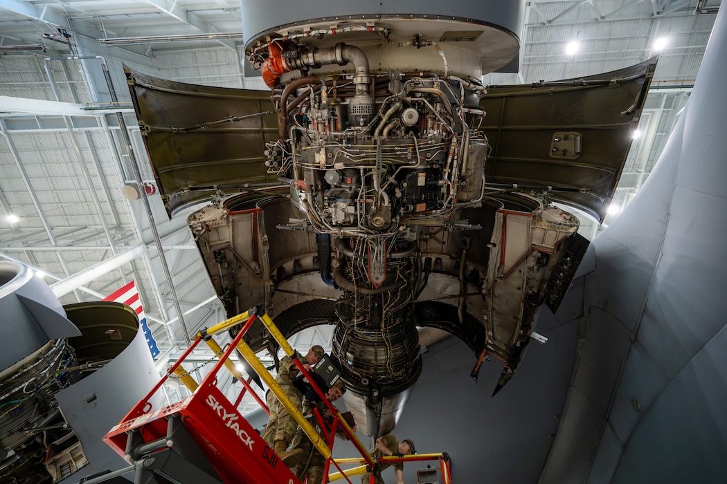 An engine is exposed with Airmen on scissor lifts around it