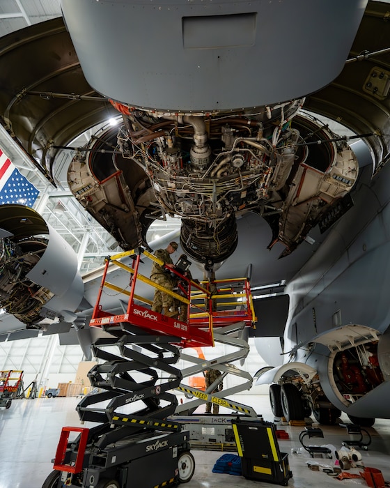 An engine is exposed with Airmen on scissor lifts around it