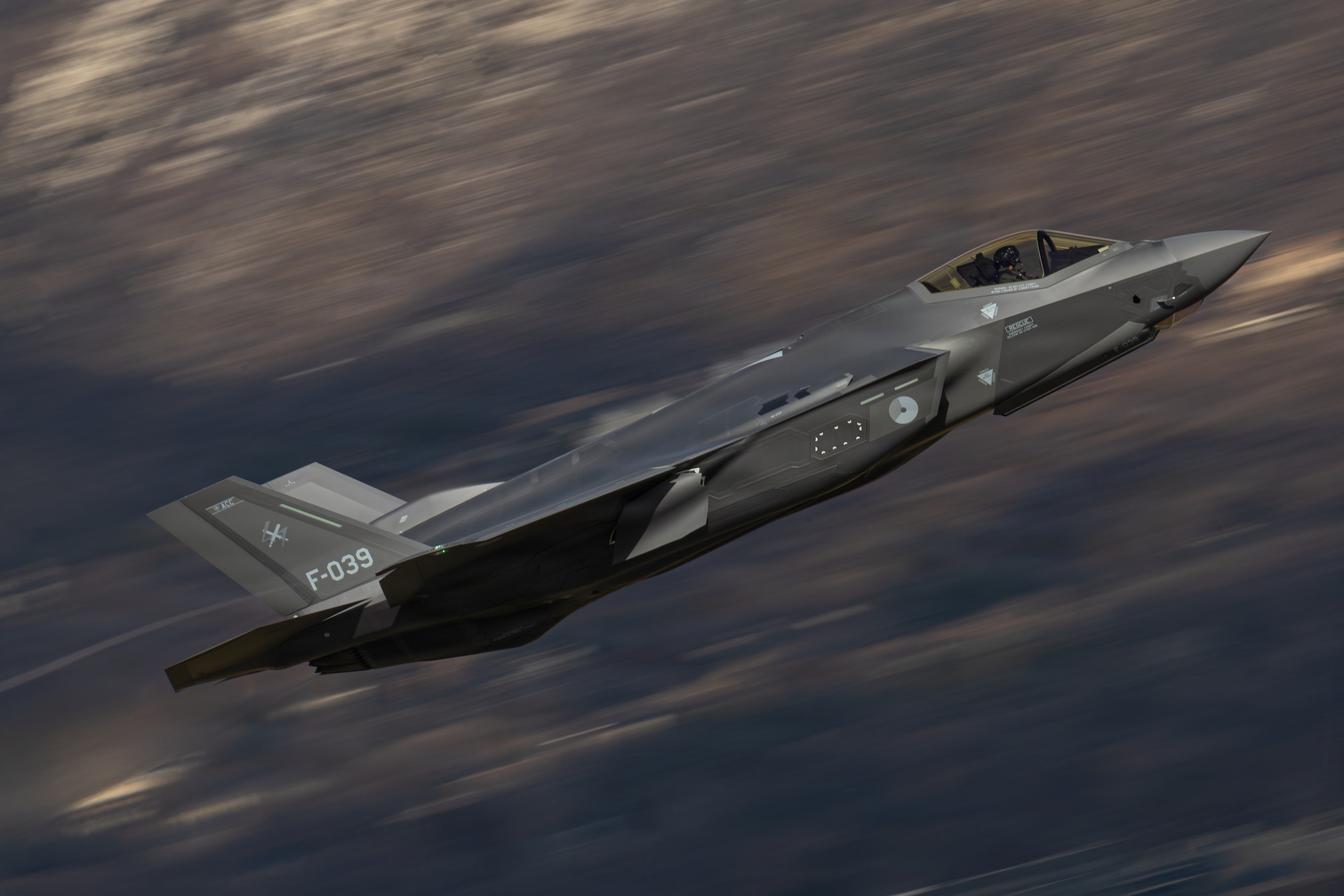 A photo of an F-35 in flight with mountains in the backgound. The mountains are blurring, demonstrating the speed with which the aircraft is flying past them.