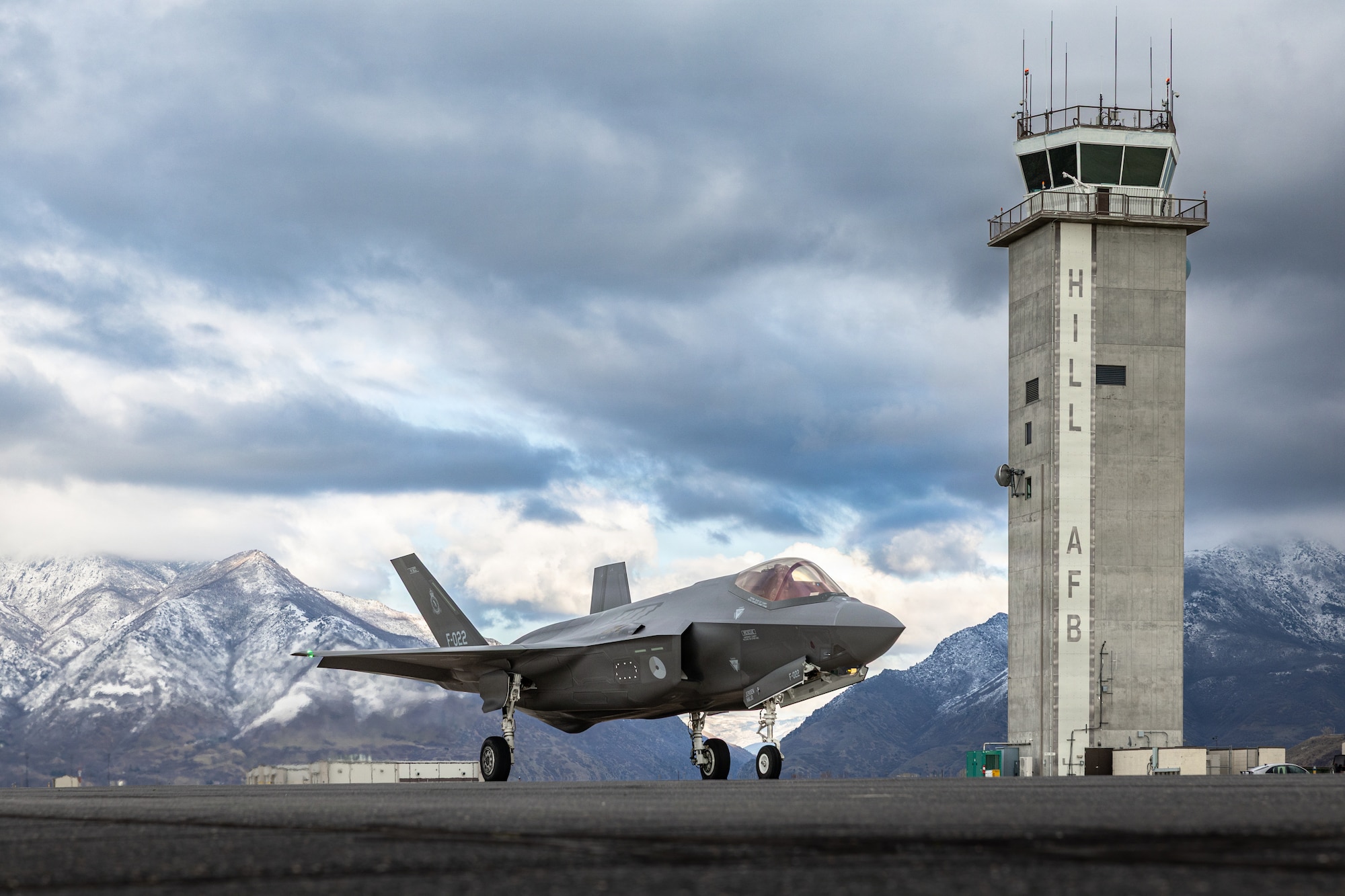 A photo of an F-35A on the flightline with an air traffic control tower and snow- capped mountains in the background