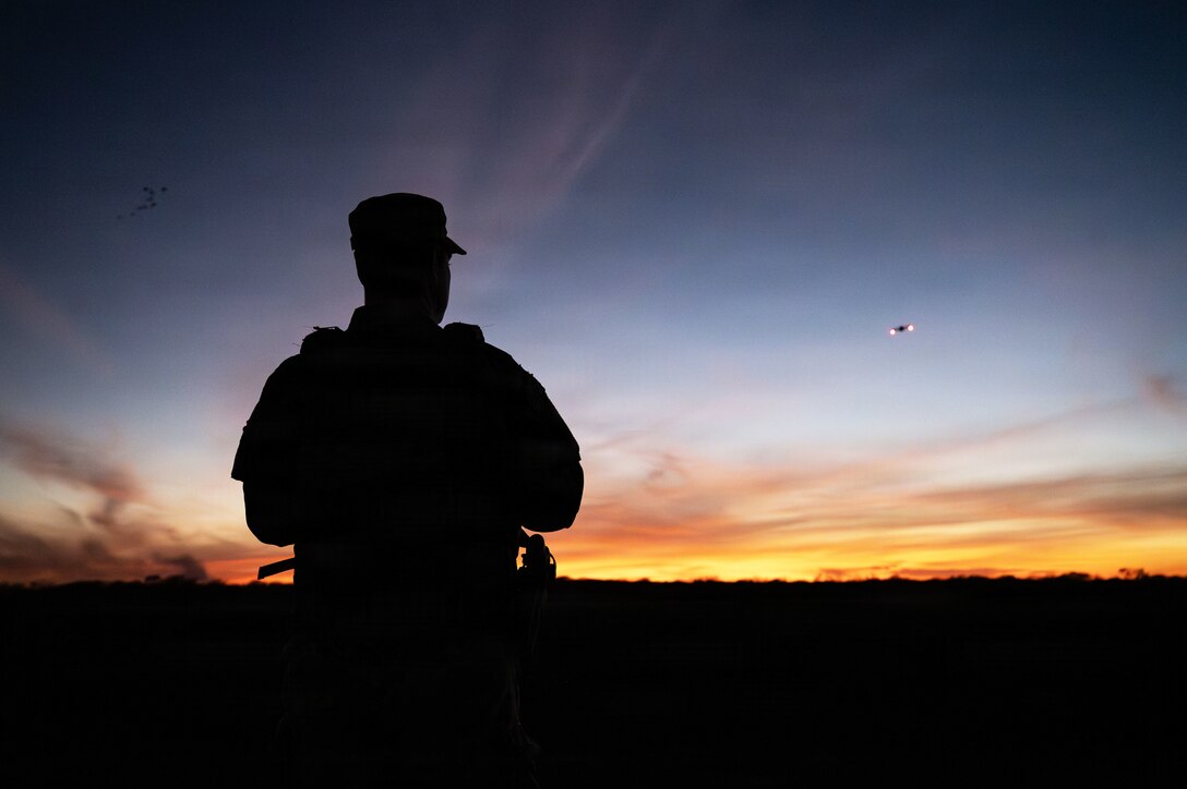 A soldier looks in the distance at a drone that is high in the sky in the distance as the sun sets.