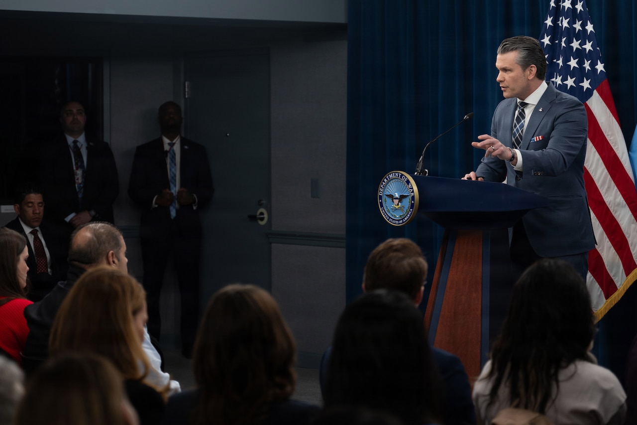 Secretary of War Pete Hegseth gestures while speaking from a lectern to a seated audience.