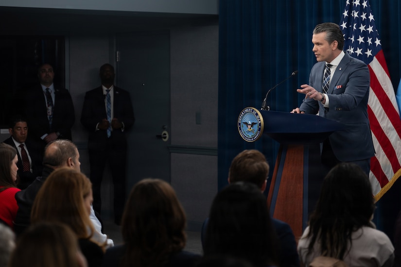 Secretary of War Pete Hegseth gestures while speaking from a lectern to a seated audience.