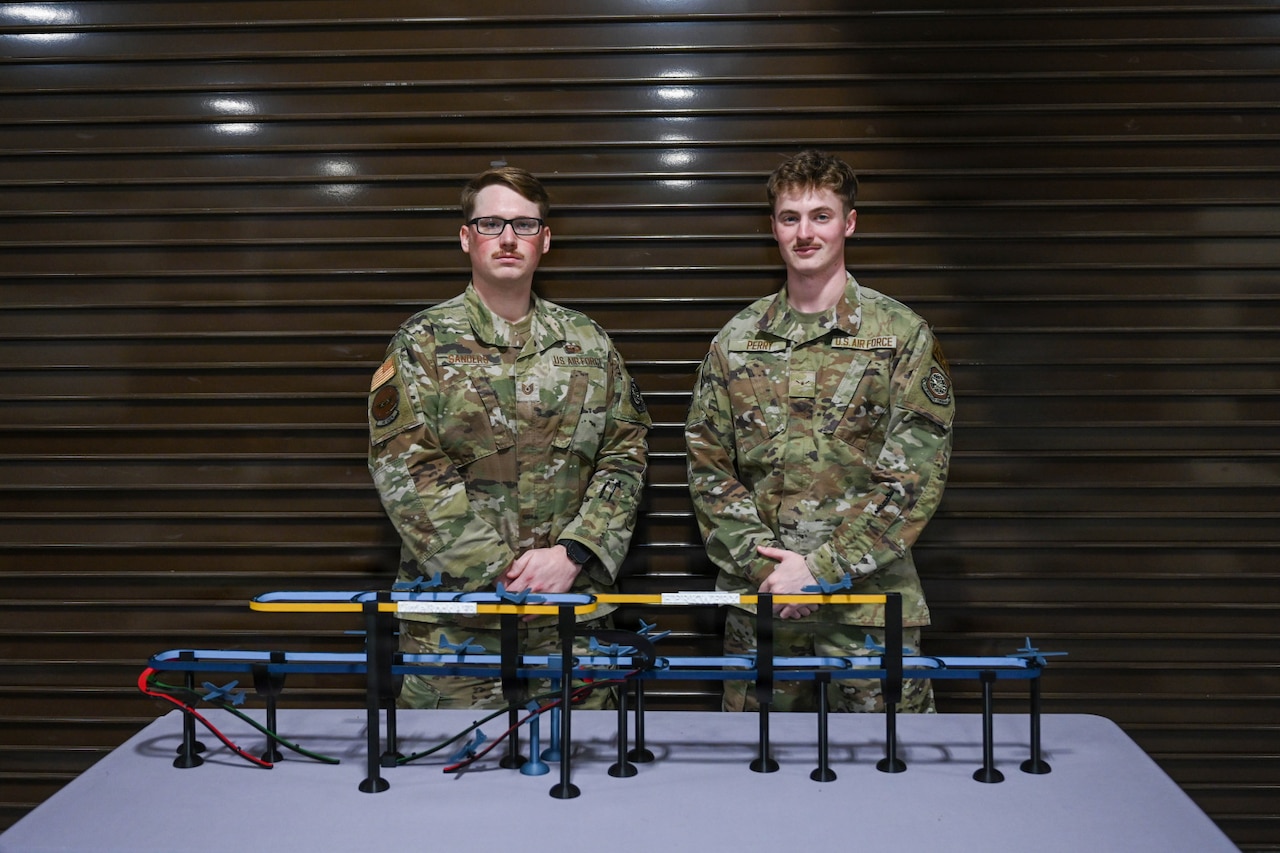 Two men in camouflage military uniforms pose for photo in front of 3D printed models of airplanes as they go through different flight patterns.