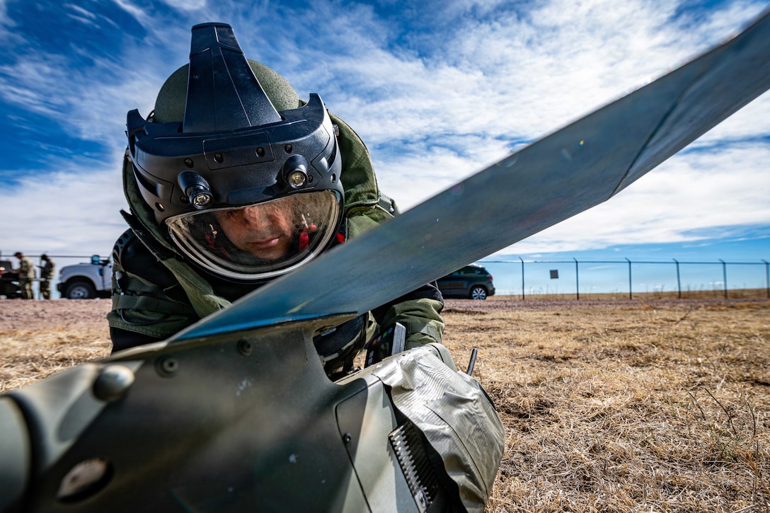 An airman in a protective suit lies in a field holding an unmanned aircraft system under a blue sky.