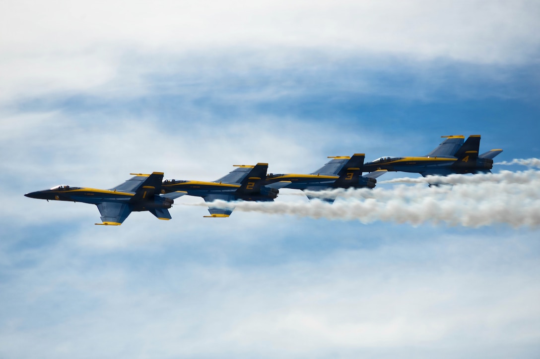 Four aircraft fly in formation in a cloudy, blue sky, leaving behind trails of smoke.