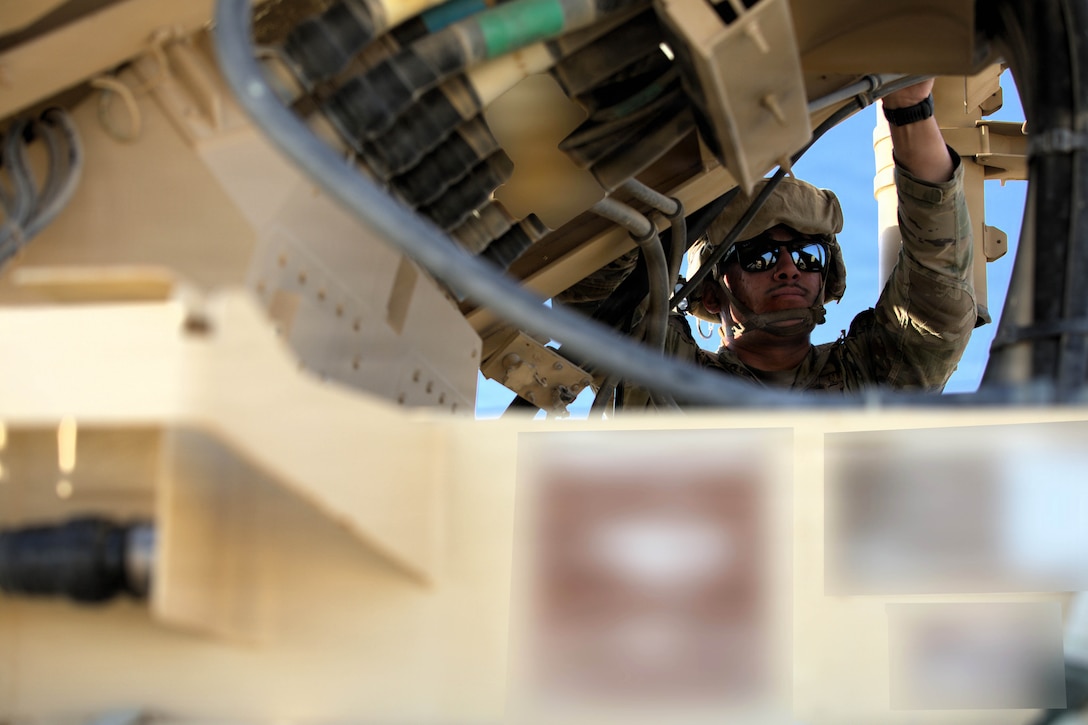 A soldier wearing a helmet and sunglasses performs maintenance on a military vehicle during the day.