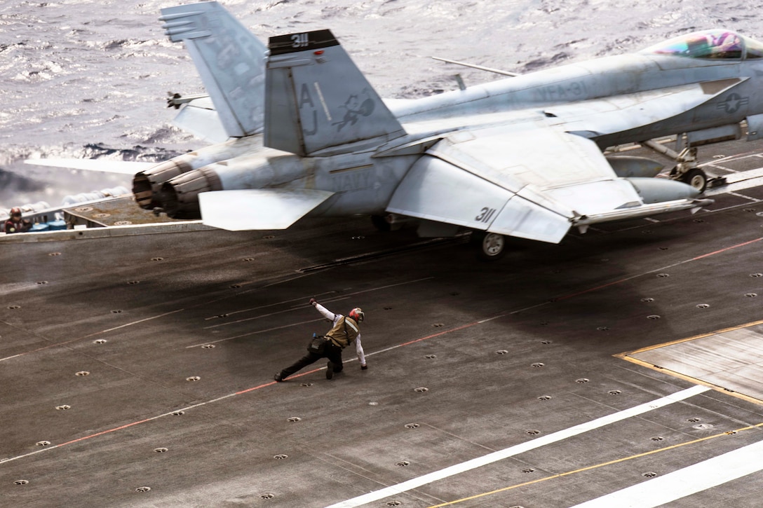 A sailor leans on the flight deck of a ship as an aircraft prepares to launch.
