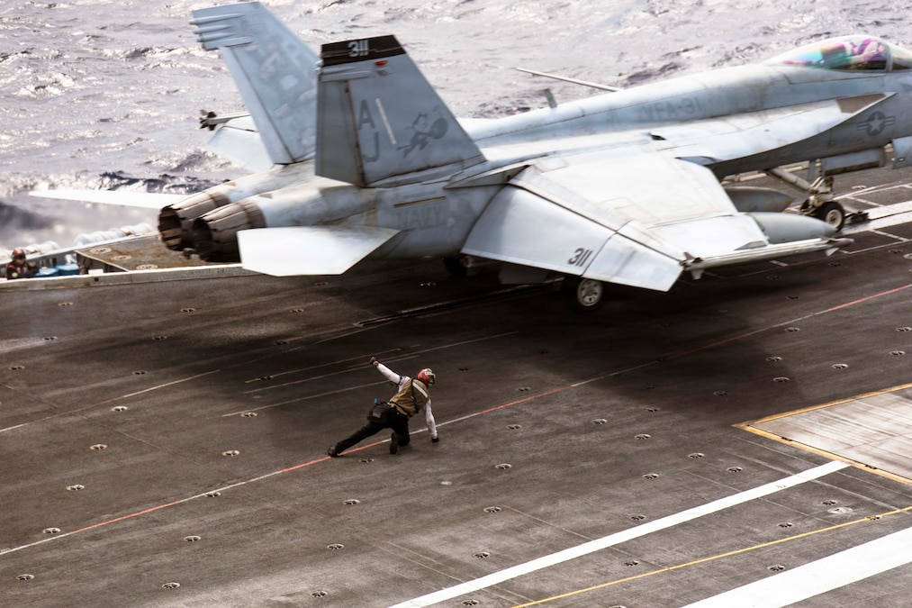 A sailor leans on the flight deck of a ship as an aircraft prepares to launch.