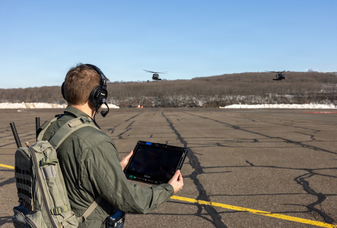 A man wearing a green flight suit, backpack and headphones, holds a tablet while watching two military helicopters in flight.