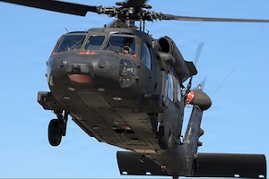 A helicopter lifts off the ground. A person wearing a helmet with a sun visor looks out the cockpit windshield.