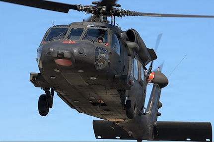 A helicopter lifts off the ground. A person wearing a helmet with a sun visor looks out the cockpit windshield.