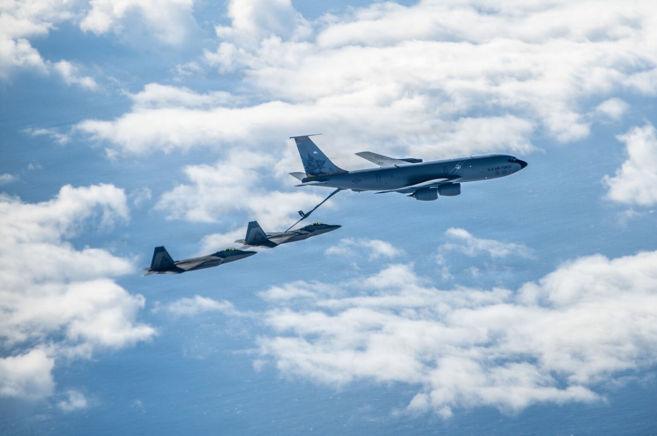Three military aircraft fly near one another. One aircraft is connected to another with a refueling boom.