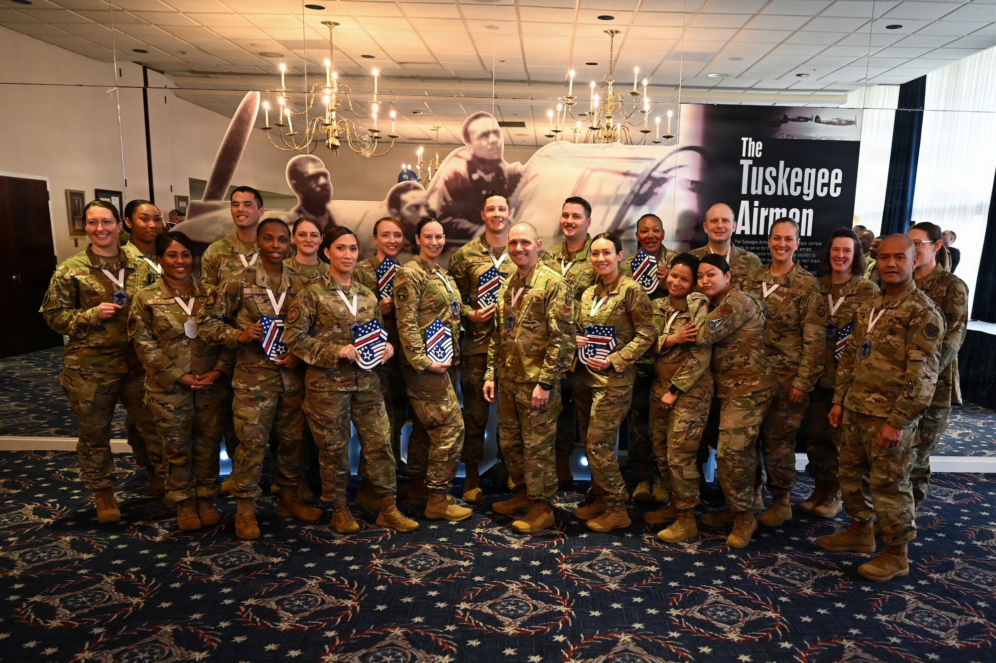 A group of 25 Airmen pose for a picture in a dimly lit ceremony room.
