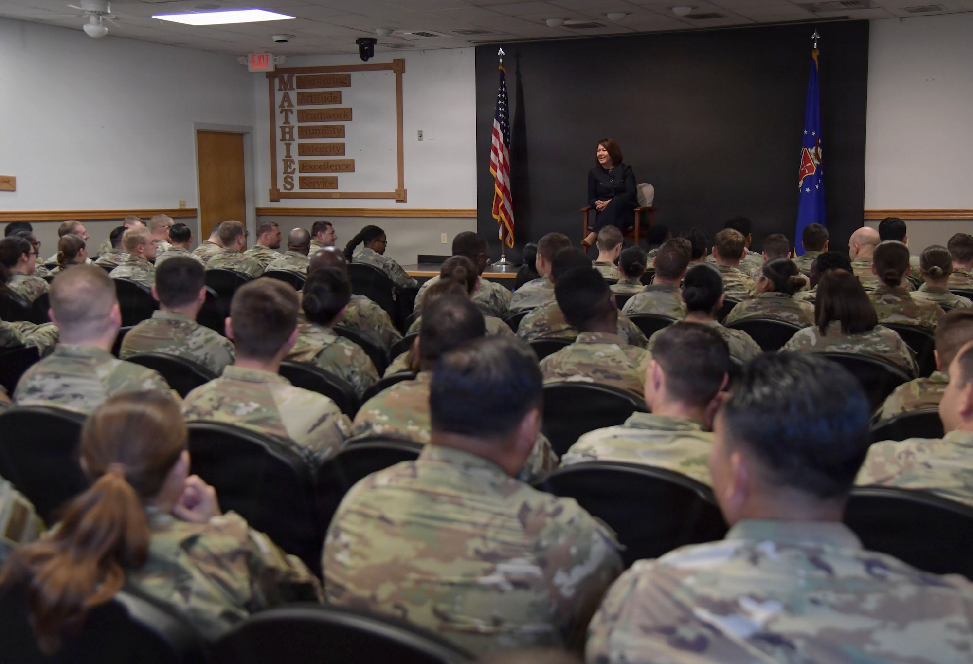 A woman speaks from a stage to a group of people in military uniforms.