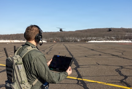 A man wearing a green flight suit, backpack and headphones, holds a tablet while watching two military helicopters in flight.
