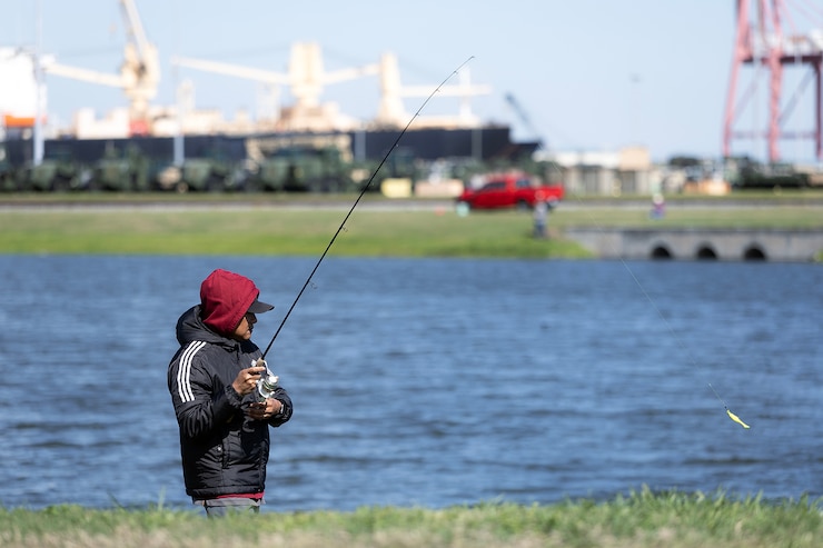 A man in a black jacket with a red hoodie fishes at a pond. 