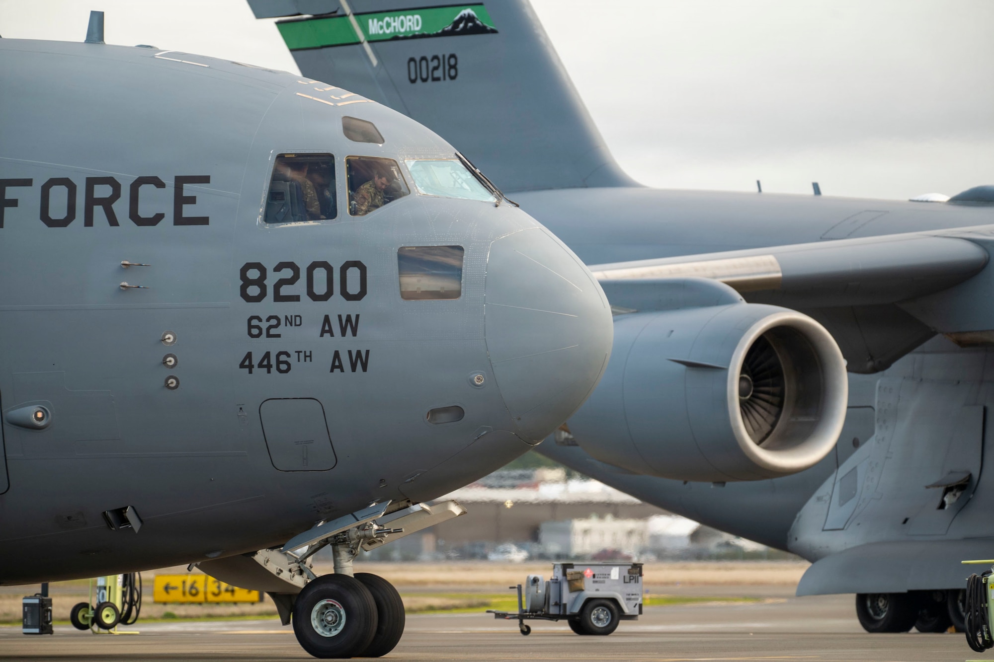 A close up of two U.S. air force cargo planes sitting on a flightline. The left side of the image only the show the front portion of one of the aircraft from the side, while the right portion shows one of the engines and the tail of the aircraft next to it