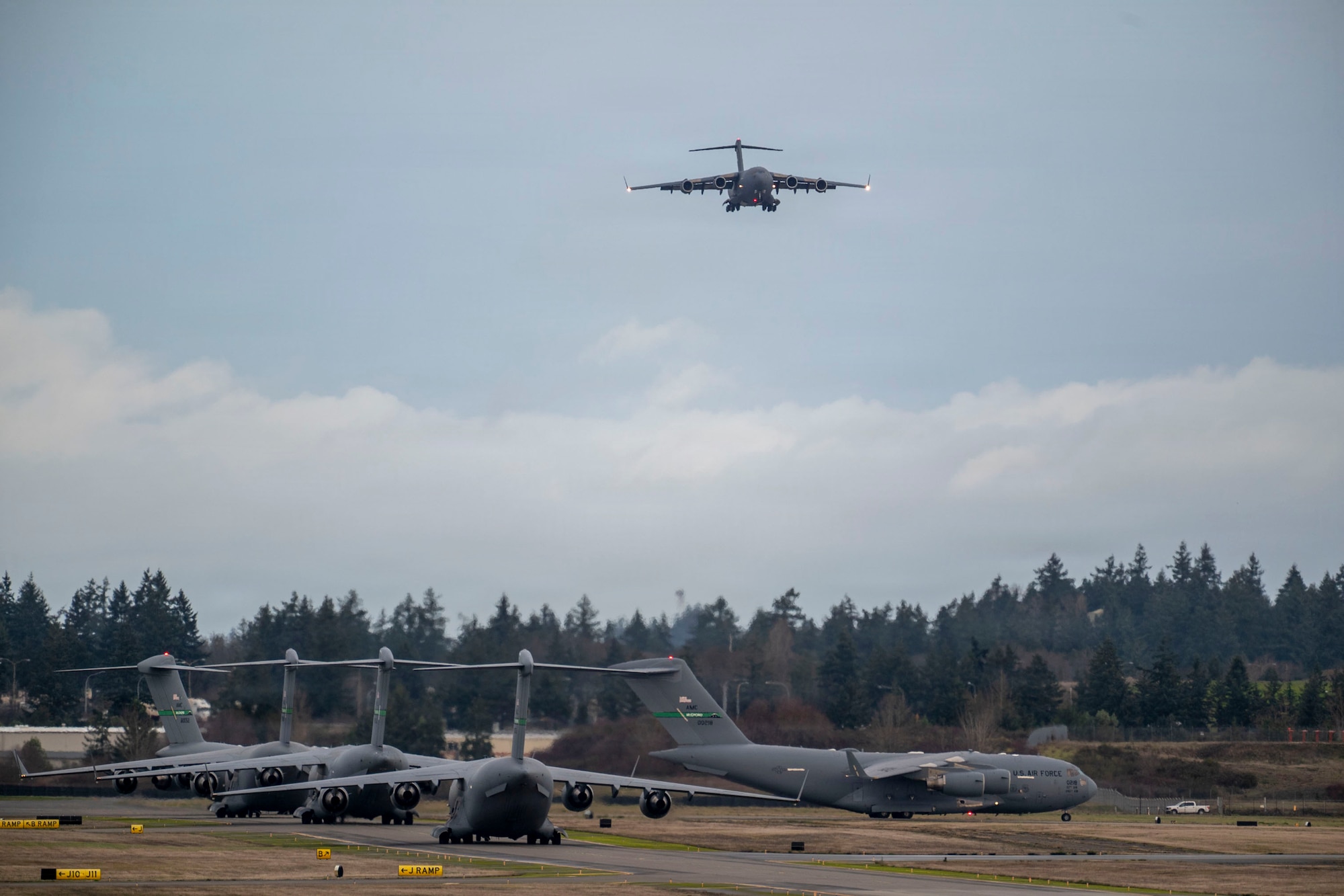 Several U.S. Air Force cargo planes taxi on a runway with 1 U.S. Air Force cargo plane flying above the rest