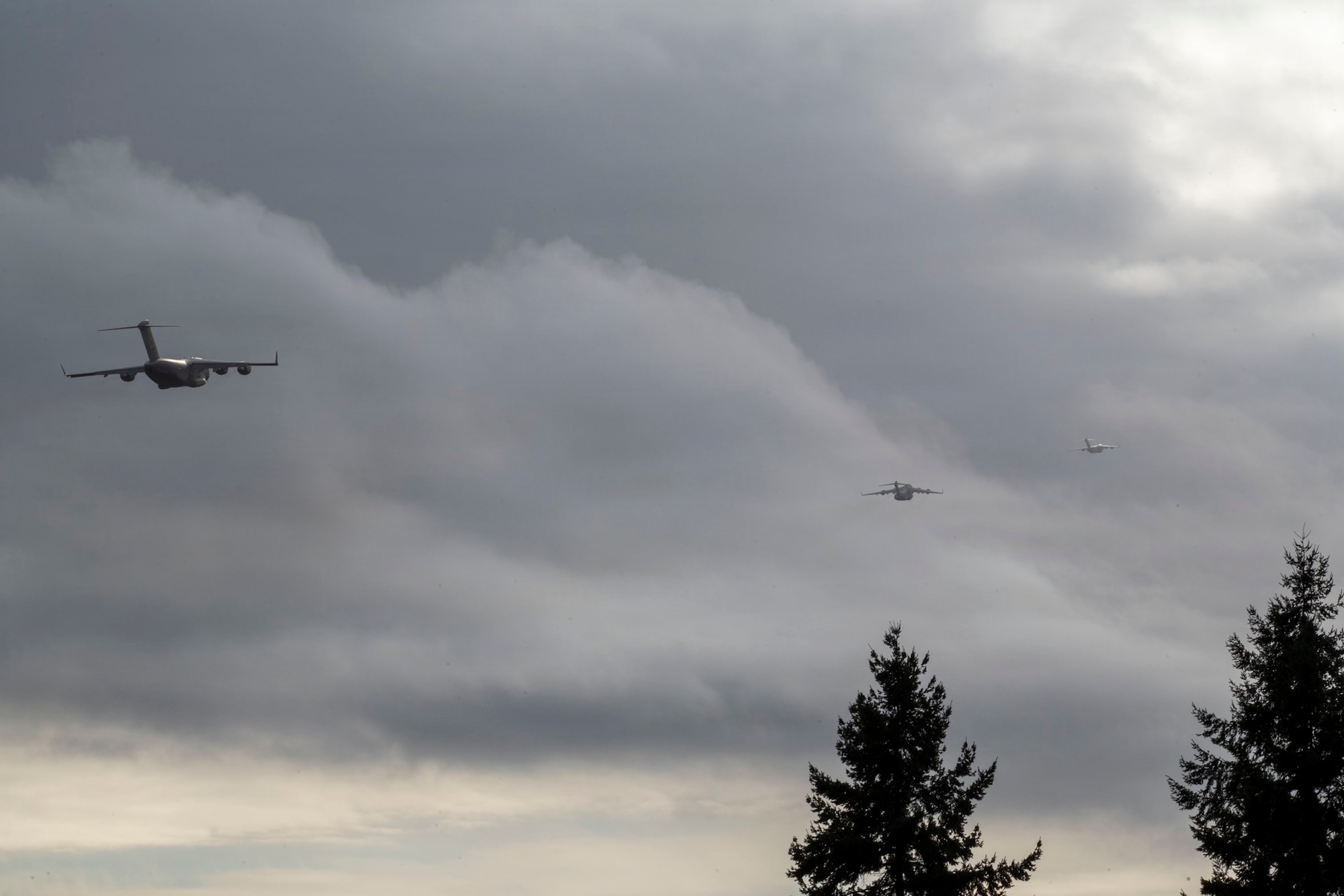 A photo of three U.S. Air Force cargo planes flying in the distance with pine trees in the foreground and a background of grey clouds
