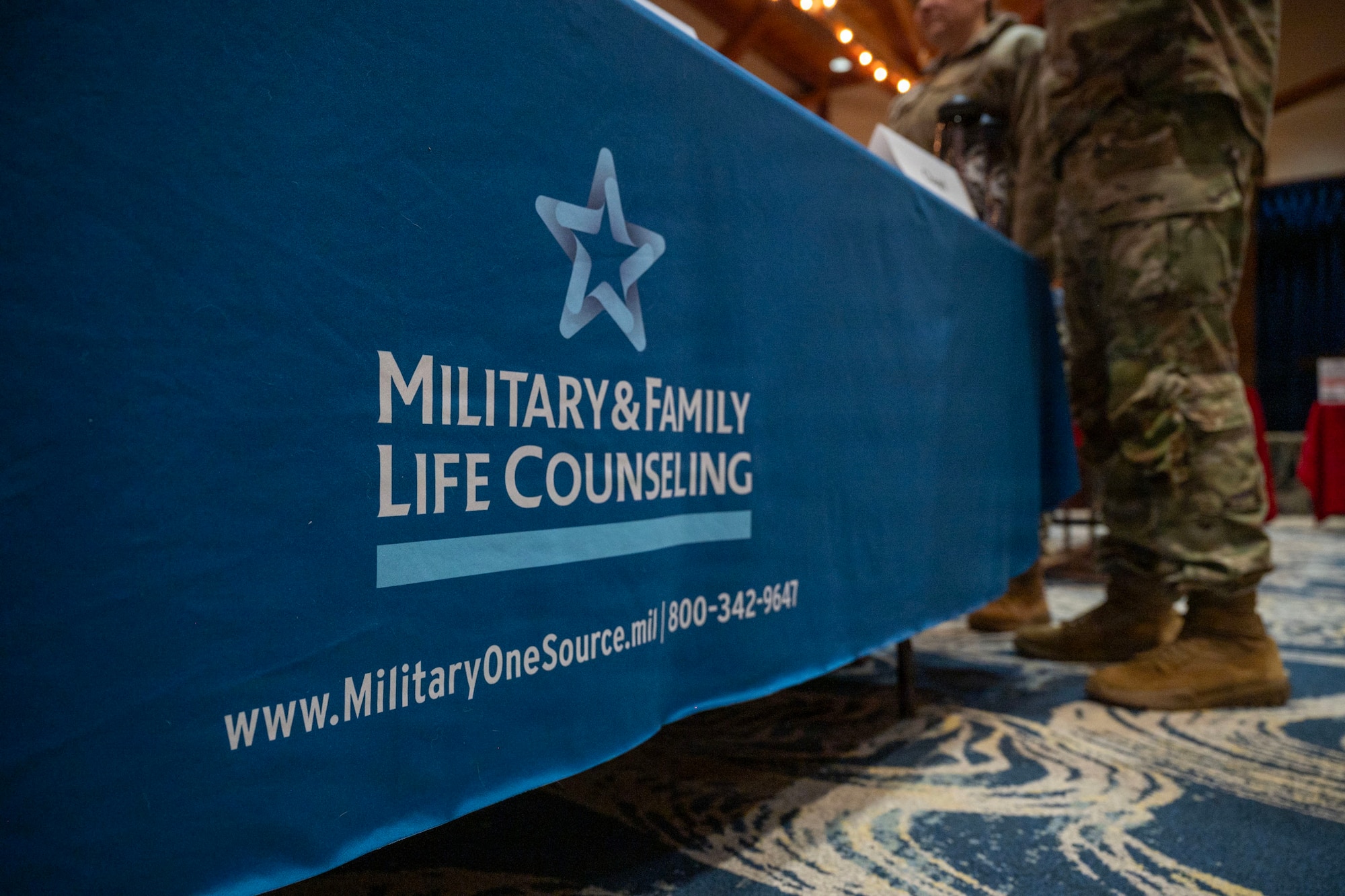 A standing booth shot from the ground displaying the text "Military and Family Life Counseling" on the front. To the right, you can see a person in military camouflage standing at the booth.