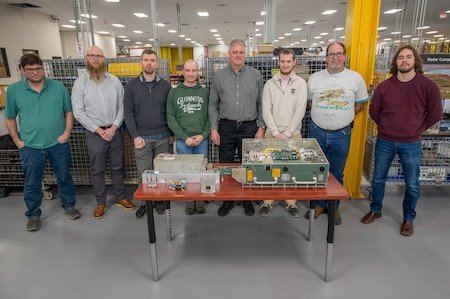 Eight people pose for a photo behind a table with pieces of a Power Amplifier Module, a piece of military equipment on it.